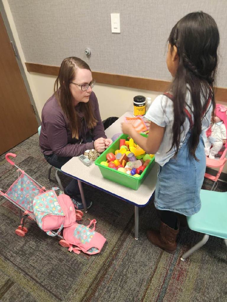 A Child Associate sits at a table with a child, various toys spread out in front of them.